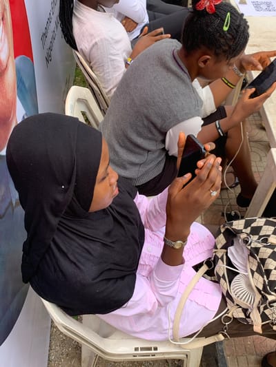 Captured from an elevated perspective, the scene depicts several individuals seated outdoors. One person wears a black hijab and a pink, long-sleeved top. Another individual holds a smartphone and has braided hair adorned with a red flower and green clips. A purse featuring a black and white checkered pattern rests on one of the chairs. The people are seated on white plastic chairs. The scene appears to be in Ilorin, Nigeria, as suggested by the text, "Nigeria: The Theme" on a graphic visible on the ground.