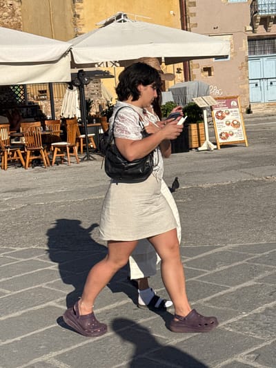 In Chania, Greece, a woman walks on a cobblestone street, illuminated by the sun. She wears a white lace top and a beige linen skirt, complemented by dark purple Crocs and a silver ankle bracelet. A black leather shoulder bag is slung across her body. She is holding a white phone in her right hand and looking at it. To her left, a person wearing a white skirt and socks with black stripes walks behind her. In the background, outdoor cafe seating with wooden chairs and tables is visible under large white umbrellas. A menu board displays food options. The buildings have weathered facades in shades of yellow and beige, with blue doors and windows. The scene is a snapshot of everyday life in a European city.