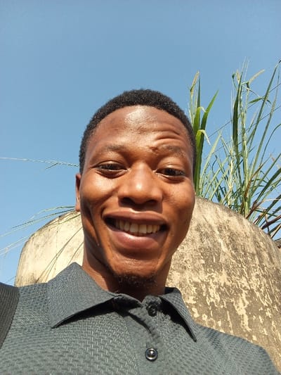 A close-up outdoor selfie features a young Black man, likely in his 20s or early 30s, smiling broadly directly into the camera. He has short dark hair and a light stubble on his chin, with a wide, open-mouthed smile that reveals his teeth, indicating happiness. He is wearing a dark grey or blue collared shirt with a subtle textured pattern, and a black strap, possibly from a bag, is visible over his left shoulder.

The image is captured from a low angle, looking upwards. Above the man's head, a clear, bright blue sky dominates, suggesting it is a sunny day, likely midday or early afternoon, given the direct light on his face. Behind him, to the right, there is a weathered, light-colored concrete or stone wall/structure with a textured surface. Tall, green grasses or reeds grow above and behind this structure, extending into the top right of the frame. The combination of the natural vegetation and the worn concrete element places the scene outdoors, possibly in a semi-urban or rural setting consistent with Aramoko-Ekiti, Nigeria. The man's joyful expression is the focal point of the sunny composition. No visible text is present in the media.