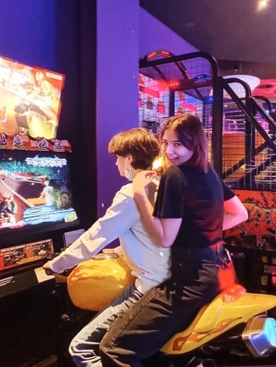 Two individuals are positioned on a yellow arcade motorcycle racing simulator, featuring red flame decals, within a dimly lit indoor entertainment venue. The person in the front, facing left, wears a light-colored long-sleeve shirt, blue jeans, and has a light-colored face mask pulled down below their chin. They are seated on the main seat, with their hands near the controls of the game. Behind them, a second person is seated on the passenger portion of the motorcycle, wearing a black short-sleeve shirt and dark jeans. This individual has shoulder-length brown hair, looks directly towards the viewer, and displays a slight smile. Their left hand rests on the front person's left shoulder.
To the left of the motorcycle, a vertical arcade screen displays game graphics. Above it, another portion of a second arcade game screen is visible. The background consists of purple-lit walls and structures. To the right, part of a basketball-themed arcade game, characterized by a net and red illuminated elements, is visible. The overall environment suggests an arcade or entertainment center setting, with artificial and colorful lighting. The specific city location of Kuyi Charhin, Uzbekistan, cannot be determined from the visual information present in the image.