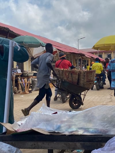 The image depicts a street scene in Kafanchan B, Nigeria. A man in a light gray shirt and dark shorts is pulling a heavily loaded wheelbarrow filled with what appears to be woven baskets. The setting is a marketplace; there are buildings with red roofing on the left, and several people are milling about, including individuals wearing yellow and red shirts. Another person is sitting on a motorcycle in the background. The area seems to be a busy marketplace with various stalls and objects visible, including umbrellas and assorted merchandise. The weather seems partly cloudy.