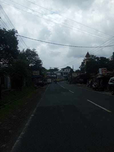 The image depicts a two-lane asphalt road under an overcast sky in Thanthirimale, Sri Lanka. Utility poles with a dense network of overhead wires span across the frame. On the left side of the road, a strip of vegetation, including trees and bushes, runs alongside a low concrete fence. Several parked vehicles, including multiple cars and a white van, are visible on the left side of the road. Further down, a yellow vehicle, possibly a bus or truck, is also present.
On the right side, a row of commercial buildings, some multi-story, lines the road. A prominent building with a light-colored facade and a dark red, conical-roofed turret structure is visible in the mid-ground. Numerous smaller roadside stalls and structures are present. A dark-colored SUV is parked partially visible in the immediate foreground on the right. Several motorcycles are parked alongside the road, with multiple individuals gathered near them, some standing and others possibly seated. Another motorcycle with a rider is visible in the distance, traveling away from the viewer. Road markings include a solid white line on the left edge and a dashed white line segment in the center, indicating two lanes. Various signs are affixed to buildings and poles on both sides of the street.