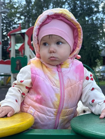 A baby in a pink hat and tie-dye jacket is positioned at an angle, likely on a playground, indicated by the presence of play equipment in the background. The baby has a thoughtful expression, looking off-center. The jacket features a zipper and is worn over a white top with cherry patterns. The setting includes greenery and elements of a playground, suggesting an outdoor location. No evidence is present that supports the location of Zav'yalovo, Russia.