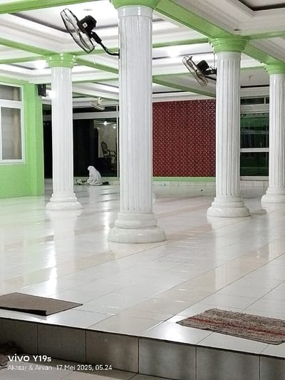 The image shows an interior space with white tiled floors and white pillars. There is one person in a white garment praying in the background, facing a wall with a red and white pattern. The ceiling has white and green trim. The image is taken in Gegesik, Indonesia, at the coordinates -6.536 latitude and 108.431 longitude.  Two ceiling fans are visible in the image. The image was captured with a vivo Y19s phone.  The time stamp on the image is 17 Mei 2023, 05:24.