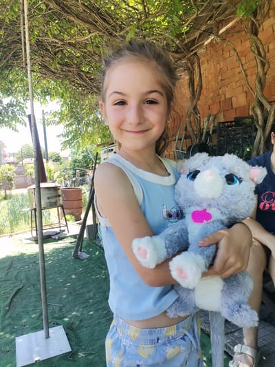 The image shows a young girl holding a stuffed gray cat. She is smiling and looking at the camera. She is wearing a blue tank top and blue shorts with a yellow floral pattern. The stuffed cat has blue eyes and a pink heart-shaped tag on its chest. The girl is standing outdoors in what appears to be a backyard. There is a brick wall behind her, and foliage from trees is overhead, creating shade. In the background, there are various objects including what looks like an outdoor oven or grill, barrels, and gardening tools. The ground is covered with green artificial turf and scattered debris. The general environment suggests a rural or semi-rural setting, potentially in Obrochishte, Bulgaria, given the presence of rustic structures and vegetation.