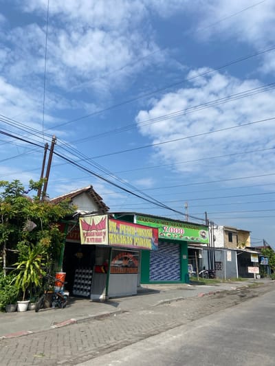 The image shows a street scene in Kecamatan Tanggulangin, Indonesia (latitude: -7.499, longitude: 112.7). There are several buildings along the street, including a small restaurant with a green sign that reads "RM. PUTRI WIKING MASAKAN PADANG". The restaurant has a white awning and a few tables and chairs outside. There is also a sign for "BAKSO & MIE AYAM PANGSIT" with a price of "7,000" displayed. In the foreground, there is a red bicycle leaning against the building. The street is paved with brick and is relatively empty of traffic. The sky is blue with some white clouds. There are multiple electrical lines crisscrossing the scene. The image was captured in the daytime.