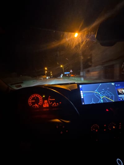 The image appears to be taken from inside a vehicle at night, potentially in Mogila, Bulgaria. The dashboard is visible, with gauges illuminated in orange light, displaying a time of 23:33 and other vehicle information. A navigation system or smartphone mounted on the dashboard displays a map. Through the windshield, street lights and signs are visible, suggesting the vehicle is on a road. The scene is dark with limited visibility, typical of night driving.