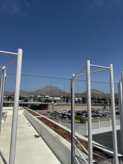 View from a high vantage point in San Bartolomé, Spain. The image features a white metal railing with a glass partition. Beyond the railing and glass is a view of buildings, a mountain, and a parking lot filled with cars. The sky is clear and blue.