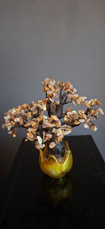 The image shows a decorative artificial bonsai tree made of polished shell pieces arranged to resemble leaves, attached to brown wire branches that sprout from a dark brown trunk. The trunk is nestled in a golden, leaf-shaped planter filled with small, shimmering particles. The planter sits on a glossy black surface, likely a table, which reflects the planter and the lower part of the tree. The background is a plain, neutral gray wall. There are no people or animals in the image, and no identifiable landmarks or street scenes. The scene is indoors and appears to be a still life arrangement. This image is not located in Orimattila, Finland.