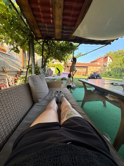 The image depicts a backyard scene in Oreshak, Bulgaria, on a sunny day. In the foreground, a person's legs are visible, resting on a wicker couch covered with a grey cushion. The person is wearing black shorts and striped socks. To the left, a portion of a concrete wall with potted plants and decorative items is visible.
In the mid-ground, two young children are present. One child, wearing a patterned dress, is climbing on a horizontal pole above a smaller child in a light-colored shirt and dark shorts. Nearby, a pink toy stroller is on the green lawn. Further back, a dark-colored ATV is parked on the grass. A large, light-colored table with chairs is set up in the yard.
The background shows houses with red tile roofs, typical of Bulgarian architecture, surrounded by greenery. Overhead, a wooden pergola covered with grapevines provides shade, with a white canopy stretched taut over part of it. The overall impression is a relaxed, family-oriented outdoor gathering in a residential area.