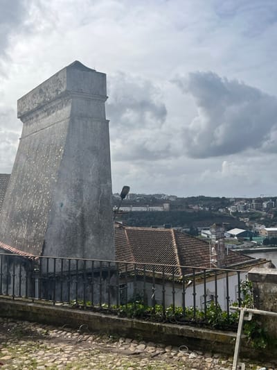 The image presents an elevated, static view of a portion of Coimbra, Portugal, under a predominantly overcast sky. In the immediate foreground, a weathered, rectangular stone pillar, exhibiting significant grey and green staining from age and environmental exposure, stands prominently on the left. The pillar tapers slightly towards a flat, minimally decorated cap.

A dark, ornate wrought-iron railing with vertical bars spans the mid-foreground, set atop a stone ledge. Below this railing, a cobblestone surface, uneven and showing signs of sparse green vegetation and debris, constitutes the ground level. Immediately behind the railing, a lower wall or parapet supports a strip of vibrant green, vine-like foliage.

Beyond the railing and foliage, a cluster of traditional buildings with distinctive terracotta-tiled roofs and light-colored walls descends the slope. A black street lamp, featuring a curved arm and an elongated lamp housing, extends upwards from behind the large stone pillar, positioned above the rooftops.

The background unfolds into a panoramic cityscape. Various structures are visible across undulating green hillsides, including a prominent, elongated, multi-story building on a distant slope. Towards the right, a body of water, identifiable as the Mondego River, is partially visible, bordered by some flat land containing what appear to be light industrial or recreational facilities. The sky is filled with voluminous grey clouds, suggesting a cloudy day. There are no individuals, animate subjects, or discernible actions depicted in the scene. The perspective suggests the photograph was taken from an elevated point within Coimbra's historic upper town, looking towards the lower city and the river.