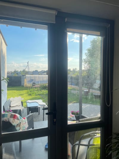 This media file is a photograph taken from inside a room, looking out through a large window. The window appears to be divided into multiple panes.

The view outside is a sunny, outdoor scene. In the upper pane, sunlight filters through green trees, casting dappled shadows on a paved patio area. A glimpse of outdoor furniture, including a chair with a bright green cushion, is visible. A small pot of pink flowers sits on a table. The sky above is blue with scattered white clouds.

The lower pane of the window shows a backyard with a neatly mown green lawn. A large, translucent greenhouse or polytunnel dominates the background, with smaller outbuildings and houses visible beyond it. A picnic table with chairs is situated near the greenhouse. There are also stacks of what appear to be building materials or large decorative items on the right side of the frame. The sky in this section is a bright, clear blue, suggesting a pleasant day.

The overall impression is one of a bright, sunny day in a residential or garden setting. The time of day appears to be late morning or early afternoon, based on the angle of the sunlight. There are no people visible in the scene. The setting seems to be rural or suburban, possibly in a region with a temperate climate. The photograph has a slightly hazy quality, which could be due to the window glass or atmospheric conditions. There is no visible text in the image.