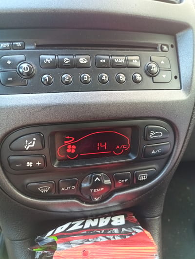The image shows a close-up view of a car's dashboard, specifically the climate control and radio console. The primary subjects are the buttons, display screen, and parts of the dashboard itself. The scene is set inside a vehicle, with the dashboard being the focal point.

The climate control display shows a temperature of "14" degrees and an "A/C" indicator. Icons on the climate control panel include symbols for airflow direction (recirculating air, face vents, and defroster for the front windshield), fan speed adjustment, and an A/C button. Above the climate controls, a car radio is visible with buttons for radio functions like RDS, TA, power, seeking channels, and preset stations numbered 1 through 6. There are also buttons for "MAN" and a music note icon, suggesting manual mode and audio controls.

The overall impression is one of a functional car interior, likely in use. There are no people visible. The lighting appears to be natural daylight, casting some shadows but not suggesting a specific time of day. The background is slightly out of focus but appears to be the interior of a car.

Visible text includes "RDS," "TA," "MAN," "A/C," "AUTO," "OFF," "TEMP," and numbers "1" through "6" on the radio and climate controls. At the bottom of the image, a partially visible wrapper for a product labeled "BANZAI" is present, suggesting that some item has been placed on or near the dashboard. The location context of Montgeron, France, does not provide any specific visual cues in the image itself. The image conveys a sense of everyday utility and technology within a vehicle.