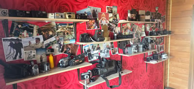 A wall covered in a vibrant red rose-patterned wallpaper displays an extensive collection of vintage cameras and photographic equipment, arranged on wooden shelves. The shelves are packed with a variety of items, including instant cameras, digital cameras, camcorders, and older film cameras. Among the cameras, numerous photographs are pinned to the wallpaper, featuring personal snapshots of people, landscapes, and possibly events. The arrangement suggests a collector's display, with items organized by shelf and seemingly by type or size. To the right of the shelves, a section of a wooden wall with horizontal planks is visible, indicating the setting might be indoors within a wooden structure or an attached area. The overall impression is one of a well-curated personal archive of photographic history. There are no people visible in the image. The location is confirmed to be in the city of Gostagayevskaya, Russia.