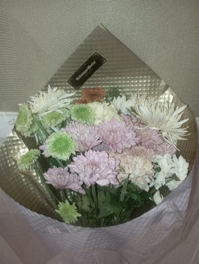 The image displays a bouquet of flowers presented in a close-up, top-down perspective within an indoor setting in Batulawang, Indonesia. The bouquet features various chrysanthemums and similar flower types, predominantly in shades of pale pink, light green, white, and a few light brown/peach tones. The flowers are wrapped in multiple layers: an innermost clear plastic sheet, followed by a textured, metallic-sheen foil with a dot pattern. A black rectangular label bearing the text "Debungaflorist" is affixed to the metallic foil. The base of the bouquet is encased in a crinkled, soft white paper or non-woven fabric wrap. The background consists of a uniformly textured, light beige or pale grey surface with subtle horizontal striations. No individuals, actions, or interactions are present in the scene.