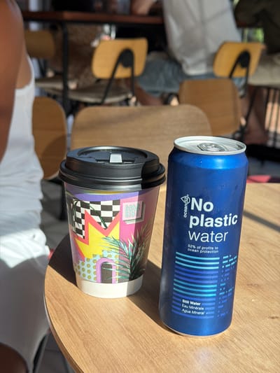 In the city of Platja d'Aro, Spain, on a wooden table, a blue can of "Ocean 52 No plastic water" stands next to a colorful disposable coffee cup. The can is condensation-covered, suggesting it is cold. The coffee cup has a black lid and a vibrant design featuring geometric patterns, palm leaves, and a stylized building. In the background, out-of-focus yellow chairs and people can be seen, indicating an outdoor or semi-outdoor seating area, likely a cafe or restaurant. A person wearing a white top is partially visible on the left side of the frame. The lighting suggests it is daytime, with shadows indicating sunlight.