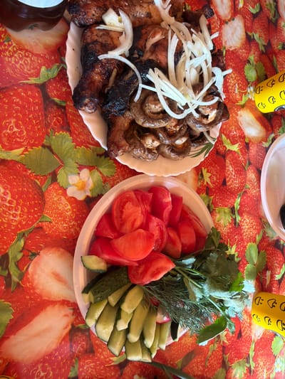 The image displays a high-angle view of a meal arranged on a table, presumably in Utash, Russia. The table is covered with a vibrant red tablecloth patterned with strawberries and white strawberry blossoms. A white, scalloped plate, centrally located in the upper portion of the frame, holds grilled meat, appearing to be chicken portions (legs and thighs) with some charring, alongside a pile of grilled mushrooms. Both the meat and mushrooms are topped with numerous thinly sliced raw white onions. Below this plate, a white plastic container is filled with a fresh salad composed of chopped red tomatoes, sliced green cucumbers, and fresh green herbs including dill and what appears to be cilantro or parsley. A dark brown bottle is partially visible in the upper left corner, and a yellow object with black decorative markings is partially visible on the right edge. No people are visible; the scene depicts food prepared for consumption.