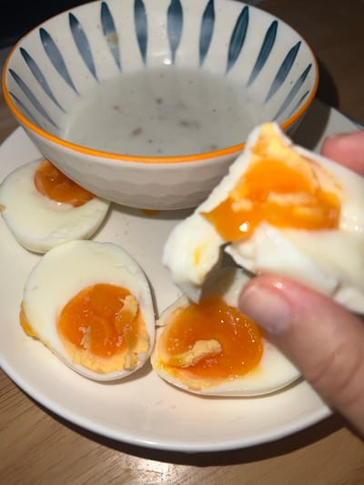 A close-up shot captures a hand holding a piece of a soft-boiled egg, revealing a bright orange, runny yolk. On a white ceramic plate, several other soft-boiled egg halves are visible, each showcasing a vibrant, semi-liquid yolk contrasting with the firm white albumen. Small grains, likely salt, are sprinkled on the surface of some yolks. To the left, a ceramic bowl with a distinctive blue-grey striped pattern and an orange rim is partially visible, containing a light greyish liquid, possibly a sauce or a thin soup. The meal is presented on a wooden surface, suggesting an indoor dining setting with artificial lighting. This meal featuring soft-boiled eggs is being consumed in the Nam Natural Heritage Area Trang, Vietnam. No text is visible in the image.