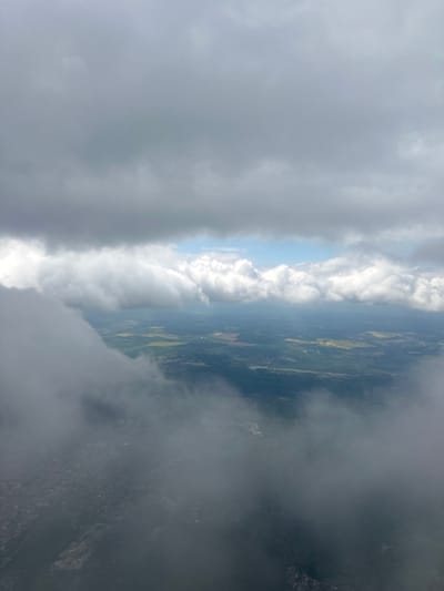 The image shows an aerial view of the landscape in the vicinity of Kladno, Czechia, taken through a cloudy sky. A section of the land is visible between the dense clouds, revealing fields, possibly agricultural lands, and a few scattered settlements. The visibility is obscured by the heavy cloud cover, which occupies a significant portion of the frame, creating a sense of a limited perspective.