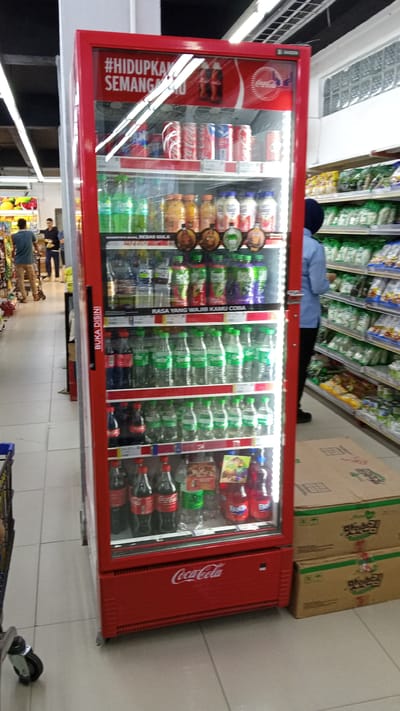 This media file captures an indoor scene within a brightly lit supermarket or convenience store. The focal point is a large, red Coca-Cola branded cooler filled with various beverages, predominantly bottled water and soda. The cooler is transparent, revealing multiple shelves stocked with drinks, including recognizable Coca-Cola products, some with green labels, and others with yellow and red hues. A prominent "#HIDUPKAN SEMANGAT" (meaning "Ignite the Spirit") slogan is visible on the top section of the cooler, alongside Coca-Cola logos. Below, text reads "BEBAS GULA" (Sugar-Free) and "RASA YANG WAJIB KAMU COBA" (A Taste You Must Try).
To the left of the cooler, a dark-haired person in a blue shirt and black pants, likely an employee, stands near a shopping cart. Another person in a blue shirt and tan pants is partially visible further back in the aisle, appearing to browse products. The floor is tiled in a light, neutral color. To the right of the cooler, stacks of cardboard boxes with Asian lettering, possibly Korean ("맛있는"), are placed on the floor.
The overall impression is a typical retail environment, emphasizing product availability and brand promotion. The lighting suggests it's daytime, and the indoor setting eliminates weather as a factor. The scene is orderly and functional, with no discernible emotions or specific events beyond regular shopping activity. The presence of multiple beverage brands suggests a well-stocked store.