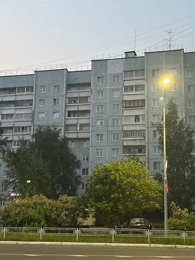The image shows a multi-story apartment building with a light blue facade. The building has numerous windows and balconies. A street lamp with a bright yellow light is visible to the right of the building. Several trees with green foliage are in front of the building, and a decorative metal fence runs along the sidewalk. A black car is parked behind the fence and trees. A Russian flag is attached to the street lamp pole. The sky above is a pale blue, suggesting it might be early morning or late evening. The scene appears to be in a residential area within the city of Ust-Ilimsk, Russia, characterized by Soviet-era architecture. There are no people or discernible actions depicted in the image.