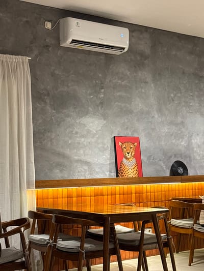 The image displays an unoccupied interior space in Patrang, Indonesia, characterized by a contemporary aesthetic. A textured grey wall, resembling polished concrete, forms the backdrop. Mounted on the upper left portion of this wall is a white GREE air conditioner unit, featuring a digital display showing "23" in blue. To the far left, white, semi-transparent curtains hang from a rod. Horizontally bisecting the mid-section of the wall is a dark brown wooden shelf. Below this shelf, a warm orange light strip illuminates a panel composed of small, rectangular orange-brown tiles arranged vertically. On the wooden shelf, a rectangular artwork depicts a cheetah wearing a prominent pearl necklace against a red background. To the right of this artwork, a black vinyl record leans upright. In the foreground, multiple dark brown wooden tables with matching chairs are arranged. Each chair is fitted with a light grey cushion secured with ties. No individuals, actions, or interactions are present in the scene.