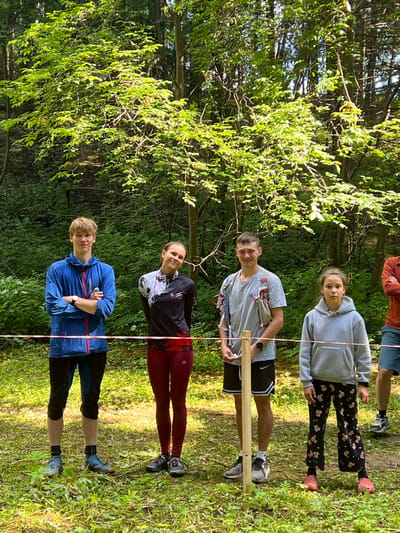Four individuals, three young males and one young female, are standing in a wooded area. They are positioned behind a red and white striped tape that runs horizontally across the frame, acting as a boundary. The individuals are dressed in athletic attire. The person on the far left is wearing a blue hooded jacket, dark pants, and athletic shoes. The second person from the left is a female wearing a patterned long-sleeved shirt with black and white accents, dark red leggings, and athletic shoes. The third person from the left is a male wearing a grey t-shirt, black shorts, and athletic shoes; he is also wearing a hydration pack. The person on the far right is a young female wearing a grey hooded sweatshirt and patterned leggings with a pinkish hue, paired with pink footwear. The background consists of dense green foliage and trees, indicating a natural, possibly park or forest setting. Sunlight filters through the leaves, creating dappled light on the ground. The ground is covered with grass and fallen leaves. The overall scene suggests an outdoor event or activity taking place in a natural environment.