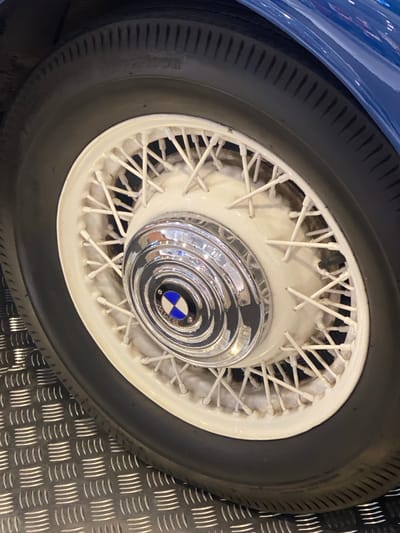 This is a close-up, eye-level shot of a spoked wheel from a vintage blue car, likely on display or in a showroom. The wheel is white with numerous thin spokes. In the center of the wheel is a chrome hubcap with a multi-layered, circular design. At the very center of the hubcap is a circular emblem with the BMW logo, featuring blue and white quadrants within a black circle, surrounded by the letters "BMW". The tire is black with a ribbed tread pattern. To the right of the wheel, a metallic floor with a diamond plate pattern is visible. The lighting is bright and artificial, suggesting an indoor setting. There is no visible text on the tire or the surrounding elements, except for the "BMW" lettering on the emblem. The image is sharp and focused on the wheel, with the car's blue bodywork visible in the upper left corner. The setting appears to be a professional display or exhibition, possibly related to automotive history or a car show, given the pristine condition of the vehicle and the specialized flooring. The location context is Łódź, Poland.