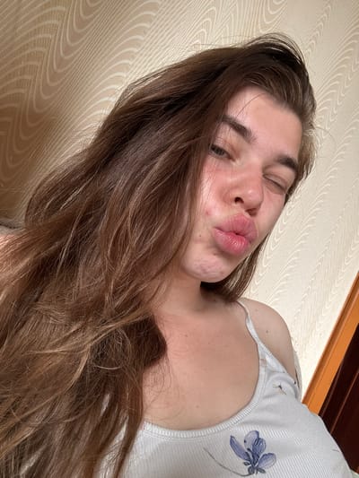 A young woman with long, brown, wavy hair is captured in a close-up, selfie-style shot. She is winking, with her lips pursed into a pout. Her face shows some minor blemishes on her chin and cheek. She is wearing a white ribbed tank top with a subtle floral pattern. The background appears to be a wall with a textured, light-colored wallpaper featuring an abstract, wavy pattern. To the right, a portion of a wooden doorframe is visible. The lighting suggests an indoor setting, possibly a bedroom, with natural light filtering in. There is no specific indication of the city of Мещёры, Russia, in the visual elements of the image.