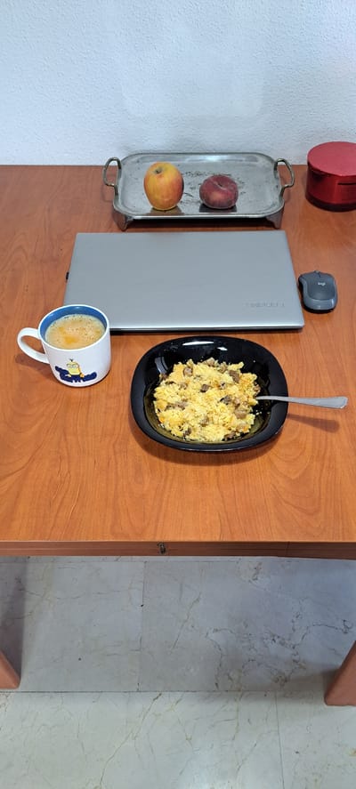 The image shows a top-down view of a wooden table. On the table, there is a silver laptop with "Lenovo" visible on its lid. Next to the laptop is a black plate filled with what appears to be scrambled eggs and pieces of meat, with a silver spoon resting in it. To the left of the laptop, there is a silver tray holding two apples, one red and one yellow. In the foreground, a white mug with a blue rim is visible, decorated with a cartoon character resembling a Minion from the Despicable Me franchise. The mug contains a frothy, light brown beverage, likely coffee. In the top left corner, a red cylindrical object is partially visible. The background shows a textured white wall and a marble floor. The scene suggests a meal is being consumed, possibly breakfast or lunch, alongside work or study indicated by the laptop. The lighting suggests it is daytime. The text "Lenovo" is visible on the laptop, and "logi" is visible on the computer mouse positioned near the laptop. The cartoon on the mug is also a notable detail. The setting appears to be indoors, perhaps a home or office.