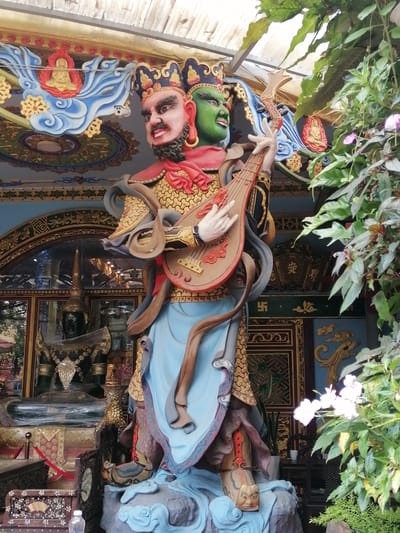 A statue of a multi-headed deity stands in front of a building in Xuân Thành, Vietnam. The statue is elaborately decorated, with a gold-colored body and a blue robe. It has two heads, one with a tan complexion and the other with a green complexion. The deity is holding a lute-like instrument. The background features ornate carvings, architectural details, and lush green foliage, suggesting a temple or shrine setting. The overall scene is vibrant and rich in cultural symbolism.