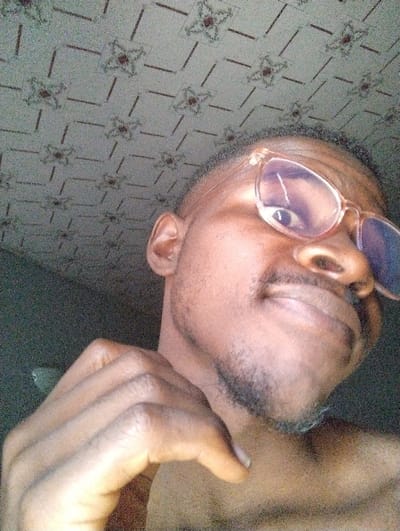 The scene is depicted as being in the city of Kaninkon, Nigeria, within an indoor setting. The image captures a close-up, upward-angled view of an adult male. The subject has a dark complexion, short dark hair, and a short beard and mustache. He is wearing clear-framed eyeglasses with reflective lenses. His right hand, with fingers curled, is positioned near his chin/neck. The individual displays a slight smile and is looking towards the upper right of the frame, indicative of a selfie. The background features a light-colored ceiling adorned with a repeating pattern of four-lobed floral designs in reddish-brown and green, interconnected by diagonal and horizontal lines. The lighting appears to be internal.