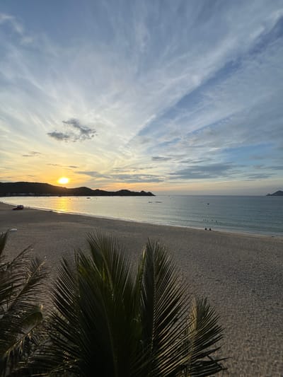 This is a photograph of a beach in Lệ Uyên, Vietnam, captured at sunset. The sun, a bright orb, is low on the horizon, casting a golden reflection on the calm ocean. The sky is a gradient of warm yellows near the sun, transitioning to soft blues and whites with wispy clouds. In the foreground, palm fronds partially obscure the view, their dark green contrasting with the warm light. The beach itself is wide and sandy, with gentle waves lapping at the shore. A line of hills borders the coastline in the distance. Several small boats are visible on the water, and a few people are scattered along the shoreline and in the ocean, some swimming and others sitting. A red vehicle is parked on the beach near the water's edge. The overall scene is one of tranquility and natural beauty.