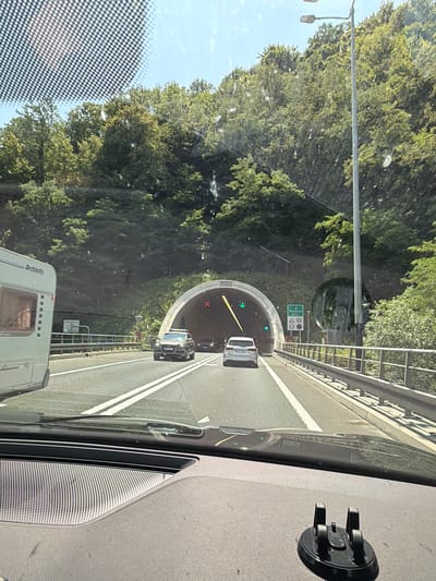 The image is taken from inside a vehicle approaching a tunnel entrance on a sunny day. The foreground shows the dashboard of the vehicle with a phone mount. A white caravan with "Dethleffs" branding is visible to the left. In front of the vehicle, a grey Audi SUV is driving in the right lane, and a white car is in the left lane, both heading towards the tunnel. The tunnel entrance is illuminated, and traffic lights and signage are visible. The tunnel is surrounded by a lush green, tree-covered hillside. A street lamp stands on the right side of the road. A green directional sign indicates "Brezovica 200 m". The road has two lanes in each direction, separated by white lines, and a metal guardrail runs along the right side. The sky is clear and blue. The location is identified as Durmanec, Croatia.