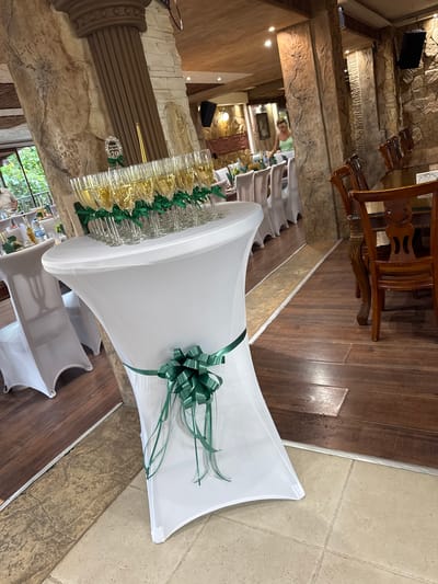The image depicts an indoor scene, likely a venue for a celebration, possibly in Sredets, Bulgaria. In the foreground, a tall, white-covered table displays a row of champagne flutes filled with a golden liquid. A decorative green ribbon bow is tied around the base of the table. Behind this table, to the left, another similar table is set with white chair covers. In the mid-ground, a woman in a light green top is visible in the background, near tables and chairs. The walls are decorated with stone and wood finishes, and there are wooden pillars. Further back, dining tables and chairs are arranged. The floor is a combination of dark wood and light-colored tile. The overall impression is of a prepared event space.