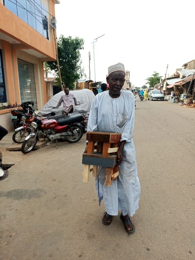 In Arawa, Nigeria, a man in a light blue and white striped traditional garment and a patterned cap walks down a paved street. He is carrying a wooden object, possibly a stool or a portable stand, made of unfinished wood. The object is composed of two pieces, one larger than the other, connected by wooden dowels. He is wearing dark sandals.
The street appears to be a marketplace, with stalls lining the right side of the road. Some stalls are covered with corrugated metal roofing and display various goods, including what appear to be pottery and other items. Several motorcycles are parked on the left side of the street, including a red and silver motorcycle in the foreground. Behind the motorcycles, a person is sitting on a white tarp-covered object, possibly a vehicle. The building on the far left has an orange facade and large, reflective windows. The sky is overcast.