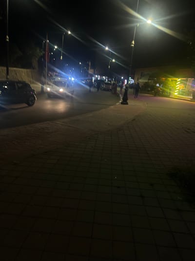The image shows a street scene in Bin El Ouidane, Morocco at night. Street lights illuminate a row of parked scooters and motorcycles along a curved sidewalk. A group of people is gathered near a brightly lit building or shop. The walkway is paved with interlocking blocks.