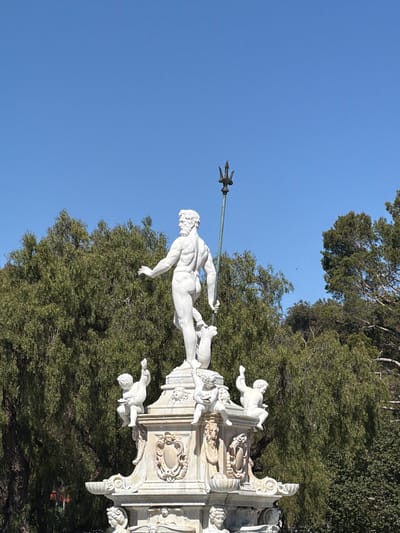The image displays the Fountain of Neptune, situated at Malaga Cove Plaza in Palos Verdes Estates, California, United States. The central subject is a white marble statue of a muscular, bearded male figure, resembling Neptune, depicted nude and holding a trident in his left hand, while his right arm is extended with an open palm. He stands atop a marine creature. Below this main figure, on an ornate multi-tiered stone fountain base, are several smaller cherub-like statues, some with raised arms. The fountain base is extensively carved with decorative motifs, including faces, escutcheons, and scrolled elements. The background consists of dense green foliage from various trees, and the sky above is clear and uniformly blue. No human figures are present, and no actions or interactions are depicted beyond the static sculptural arrangement.