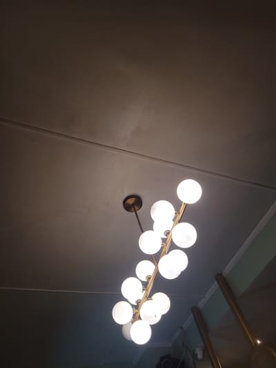 An upward-angle, low-light photograph shows a modern brass chandelier with multiple globe lights hanging from a ceiling in Manali, India. The ceiling appears to be textured and off-white, with two horizontal lines visible, suggesting a dropped or paneled ceiling. The chandelier itself is the primary subject, with its brass structure supporting a cluster of glowing white spherical bulbs arranged in an organic, branch-like pattern. Some of the bulbs are brighter than others, indicating variations in illumination and possibly their distance from the camera. The lower portion of the image reveals hints of a room, including the top of some gold-colored poles, a light green wall, and white decorative molding. The overall atmosphere is dimly lit, with the chandelier being the main source of light in the captured frame.