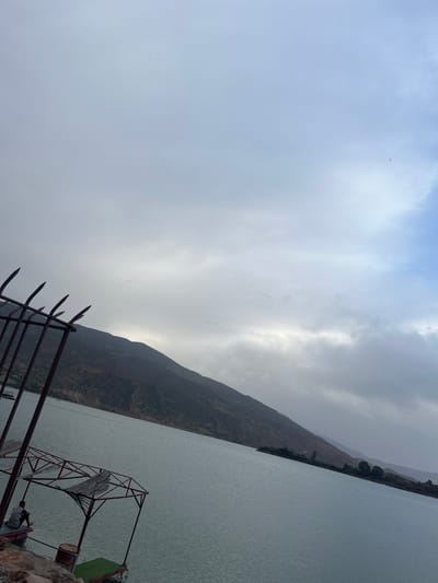 The image, oriented correctly, presents a vast body of water, likely a reservoir or lake, bordered by towering mountains under a predominantly overcast sky. The water's surface is a tranquil, light greenish-grey, exhibiting subtle ripples across its expanse.
In the immediate foreground, to the left, is a rusty metal fence or barrier topped with sharp spikes. Further to the right, a more elaborate red metal structure, possibly a gantry, part of a boat dock, or a viewing platform, extends towards the water. A person, dressed in light-colored attire, is partially visible, sitting or standing near the base of this red structure.
The distant mountains lining the far bank are dark and rugged, with some visible vegetation, and their upper slopes are shrouded in low-hanging clouds. The sky is characterized by thick, grey and white clouds, with brighter areas indicating where the sun attempts to penetrate, casting a soft, diffused light across the entire scene. The weather appears cloudy and muted, contributing to a serene yet somewhat somber atmosphere. No specific activity is depicted, nor is any text visible. The scene is set in Bin El Ouidane, Morocco, showcasing the dramatic landscape of the region, likely around the Bin El Ouidane Dam.