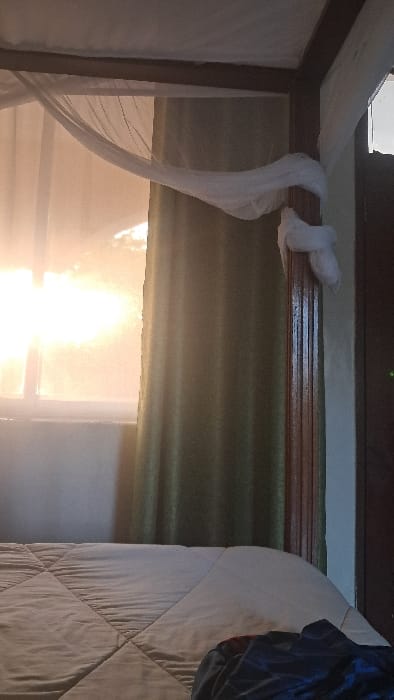 The image depicts an indoor scene, likely a bedroom, characterized by soft lighting and various textures. In the upper half, a dark brown wooden beam or frame runs horizontally, to which a piece of white, sheer fabric is loosely tied. This fabric, resembling mosquito netting or a decorative bed canopy, hangs gracefully down the left side of the frame, partially obscuring the view.
Below the wooden beam, a dark green, horizontally pleated fabric, possibly a curtain or roller blind, covers a portion of a window. An intense, bright light, appearing almost white or yellow, streams in from the window, creating a strong glare that dominates the lower center of the image. This bright light suggests it is daytime, possibly with direct sunlight.
On the far right side of the image, a large section of a bed is visible, covered with a light-colored, likely white or off-white, quilted comforter or duvet. The quilting pattern consists of diagonal lines forming a diamond or square grid. Resting on the comforter in the upper right corner is a crumpled piece of dark fabric, possibly clothing, with hints of blue, black, and red. The room appears to be softly lit apart from the direct glare from the window, highlighting the textures of the fabrics and the wooden structure. No text is visible in the image.