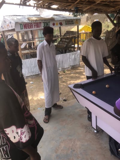 The image captures an outdoor scene in Dabawa, Nigeria, featuring five young males under a rudimentary shelter made of wooden poles and branches. In the foreground to the right, a white-framed pool table with a dark blue felt surface is present, displaying two orange-striped pool balls. A male in a white t-shirt and dark trousers stands with his hands resting on the table, facing left. To his left, another male wearing a long white tunic, dark trousers, a maroon cap, and flip-flops stands with his arms clasped behind his back, observing the table. To the far left, parts of three other males are visible; one in a black graphic t-shirt is partially seen from behind, and two others are partially visible to his left, seemingly observing the activity around the pool table. The ground consists of a concrete slab in the immediate foreground and a dirt/sandy area with scattered debris further back. In the background, a structure with corrugated metal walls and roof is visible, bearing a sign that reads "D MAGA FRIUT JOINT" (sic) with illustrations of an apple, watermelon, bananas, and two phone numbers (0706584165, 0802932567). Beyond this structure, a yellow corrugated metal building is partially visible.