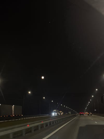 The image depicts a nighttime view of a section of the bridge spanning the Oka River in Tatarintsevo, Russia. The right side of the frame features the guardrail of the bridge with closely spaced yellow lights. Additional lights illuminate the dark expanse of the river from bridge supports. A line of lights stretches away from the viewer towards the horizon in the background. The left side of the image shows the dark expanse of the river.