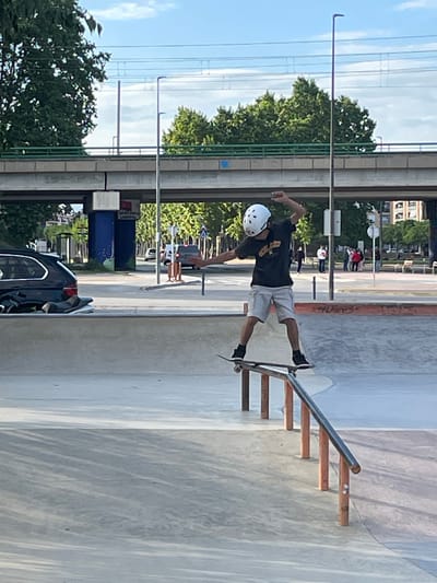 This image depicts a skateboarder performing a trick on a metal grind rail in an outdoor skate park. The scene is set on a bright, sunny day, likely in the late morning or early afternoon, under a clear blue sky with a few wispy clouds.
The main subject is a skateboarder, wearing a white helmet, a dark t-shirt with a light-colored graphic design, light-colored shorts, and dark shoes. They are in mid-action, with their skateboard balanced on an inclined metal rail that is supported by wooden steps. The skateboarder’s body is angled, and one arm is extended for balance, conveying focus and control during the trick.
The background reveals an urban or suburban environment. A line of lush green trees borders the skate park, behind which an elevated road or bridge structure with its supporting pillars and overhead power lines is visible. Distant buildings can be seen under the bright sky. On the ground level, a dark-colored car is partially visible, parked near a blue public trash or recycling bin. Faint text on the bin, possibly in Catalan, includes phrases like "PURA I SIMPLE" and "RECYCLE." The skate park itself features smooth concrete surfaces and hints of other ramps or bowls. The overall impression is one of active recreation in a sunny public space in Santa Perpètua de Mogoda, Spain.