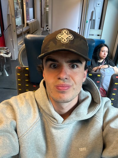 The image, rotated 90 degrees clockwise, presents a close-up selfie of a man inside what appears to be a train carriage. The man, with dark hair and light stubble, looks directly at the camera with wide, expressive eyes and slightly parted lips, conveying a playful or surprised emotion. He is wearing a camouflage-patterned baseball cap featuring a distinctive white, stylized quatrefoil or intertwined knot logo on the front. He is also dressed in a light grey hooded sweatshirt, on which the white embroidered text "6pm" is visible on the right side.
In the mid-ground, a woman with long, dark hair is seen resting or sleeping in a train seat, her face partially visible and her expression relaxed. She is wearing a dark top, and a light-colored bag rests beside her. The train seats in the background and for the woman are upholstered in a dark fabric with a repeating pattern of small red and yellow rectangular dots. The interior of the train is visible, including light-colored wall panels, reflective surfaces, and a metal luggage rack above the windows. The scene depicts two individuals traveling, with one actively engaging the camera and the other at rest. The setting suggests a public transport environment, likely a train journey in daylight hours.