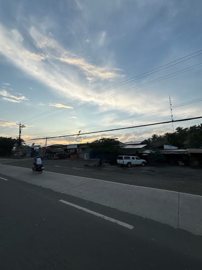 In Carmen, Philippines, a rural road runs alongside a dirt lot filled with various structures, possibly small businesses. The sky is a soft blue with wispy white clouds, suggesting either dawn or dusk. A single motorcycle with a rider is traveling on the road. A white pickup truck with a camper shell is parked in the dirt lot. In the background, a large construction crane is visible, and tall antennas dot the landscape, indicating some level of infrastructure. The scene depicts a slice of everyday life in a developing area, with a mix of transportation, commerce, and communication technology under a serene sky.