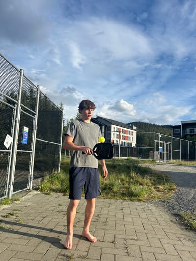 A young man is standing barefoot on a paved walkway in Whistler, Canada. He is holding a black pickleball paddle in his right hand and a yellow pickleball is suspended near the paddle. He is wearing a gray t-shirt and navy blue shorts. To his left is a chain-link fence, and beyond that, grass and trees. In the background are buildings and a forested hillside. The sky is blue with scattered clouds. A pickleball court can be seen in the background near the buildings.