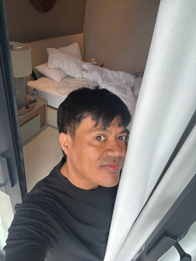 An eye-level shot of a man with dark hair, wearing a black long-sleeved shirt, looking directly at the camera. Behind him, a bright white curtain is partially visible. The background reveals a glimpse of a cluttered bedroom: an unmade bed with white linens, a bedside table with a lamp, and a section of carpet. The picture is likely taken through a window or a glass door.