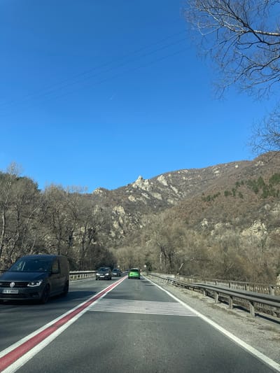 The image depicts a mountain road near the city of Poleto, Bulgaria, on a clear, sunny day. The scene is dominated by a two-lane asphalt road in the foreground, marked with a solid red line and white solid line on the left edge, a white dashed line separating lanes, and white horizontal hatch markings on the right side of the left lane. A dark gray Volkswagen Caddy van with Bulgarian license plate "PK 9144KB" is positioned in the left lane, heading away from the viewer. Further down the road in the right lane, two dark sedans and a bright green car are also traveling in the same direction.
To the right of the road, a metal guardrail extends into the distance, separating the road from a sloping embankment covered in bare deciduous trees and dry vegetation. On the left side of the road, similar bare deciduous trees line the roadside, transitioning into dense forest further back. The background is dominated by a rugged mountain range, characterized by rocky outcrops and slopes covered with a mixture of bare deciduous trees and scattered evergreen patches, indicative of a late winter or early spring season.
The sky above is bright blue and clear, with three thin, dark power lines visible horizontally in the upper left quadrant. A faint white contrail from an aircraft is discernable in the upper center-left of the sky. No individuals are visible in the scene. The overall environment suggests a rural mountain pass.