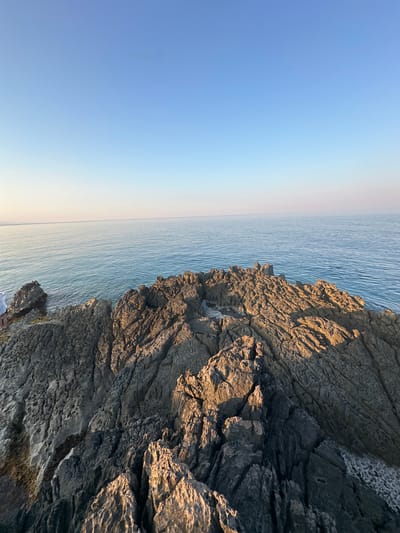 The image depicts a coastal scene in Kovačko Polje, Montenegro. In the foreground, rugged, weathered rocks jut out into the sea. The rocks are textured with cracks and crevices, with some areas illuminated by the warm light of the setting sun, casting long shadows. The sea is a calm, deep blue, with gentle ripples on its surface. The horizon line is sharp, separating the water from a clear, pale blue sky that fades to a lighter hue near the horizon. A person, partially visible on the far left edge of the frame, stands on the rocks, appearing to observe the sea.
